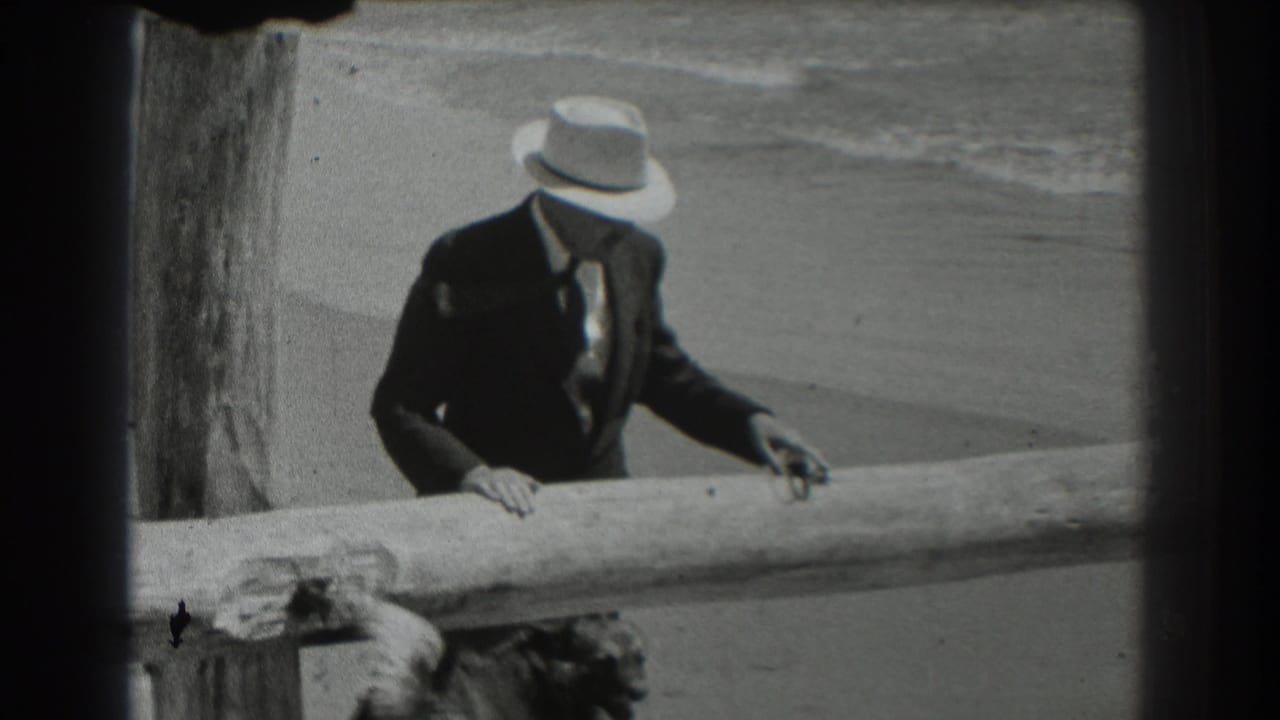 Man in a fedora leans over a wooden beach railing with surf behind him.