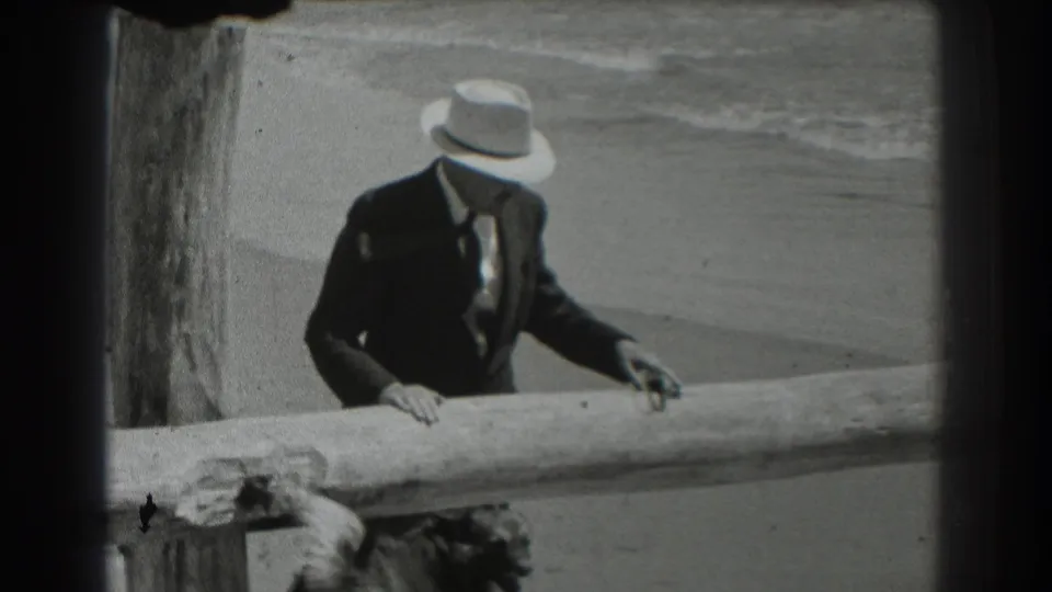 In 1939 San Francisco beach footage, a suited man in a light fedora leans toward a dog partly visible below a wooden railing.