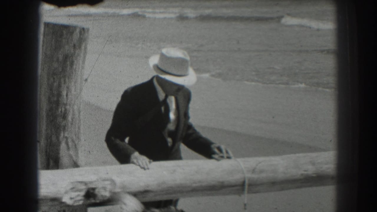 Fedora-wearing man reaches toward a rope or leash at a beach railing.