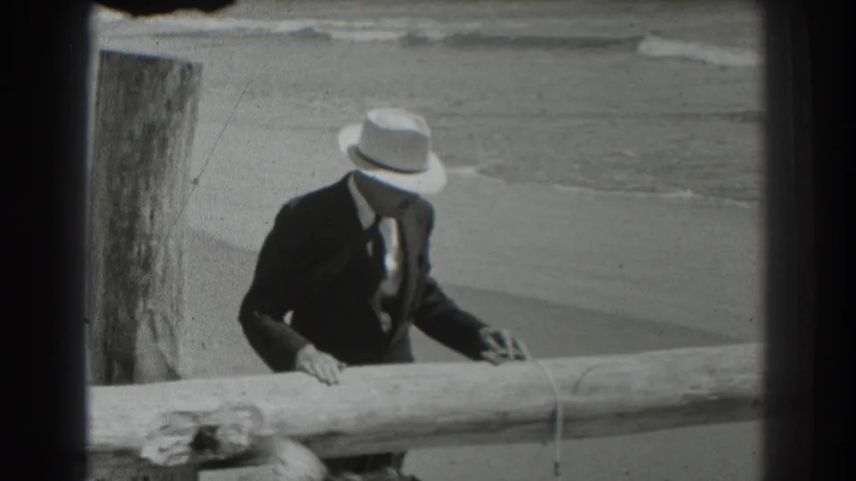 The man stands at the San Francisco beach railing, reaching toward a leash or rope while the shoreline remains in the background.