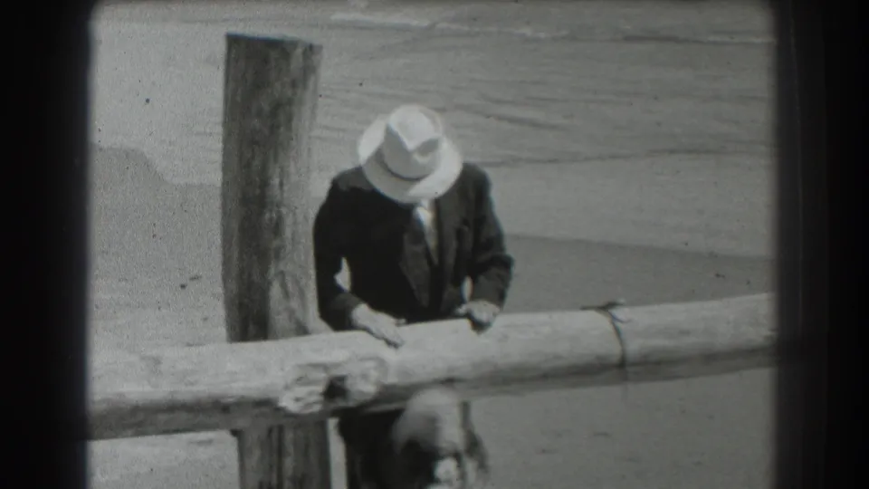 A suited man in a fedora looks down over the wooden railing toward a partially visible dog in this 1939 San Francisco beach scene.