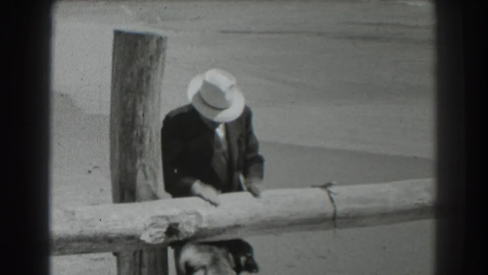 1939 San Francisco 8mm frame showing a suited man in a light fedora leaning over a wooden fence toward a dog.