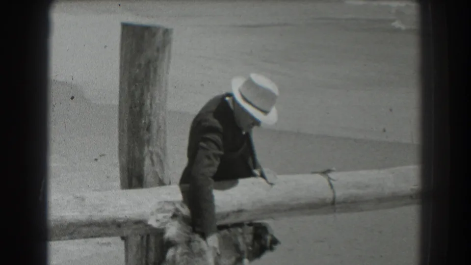 1939 San Francisco home-movie frame of a suited man reaching over a wooden fence toward a dog near the rail.