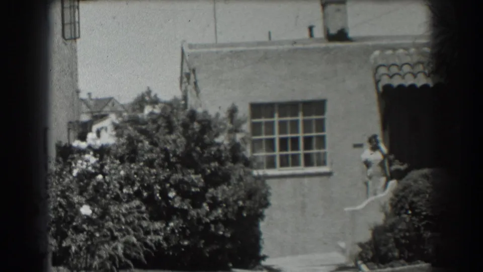 1939 San Francisco 8mm frame showing a stucco house exterior, shrubs, a window, and a person standing near an entrance.