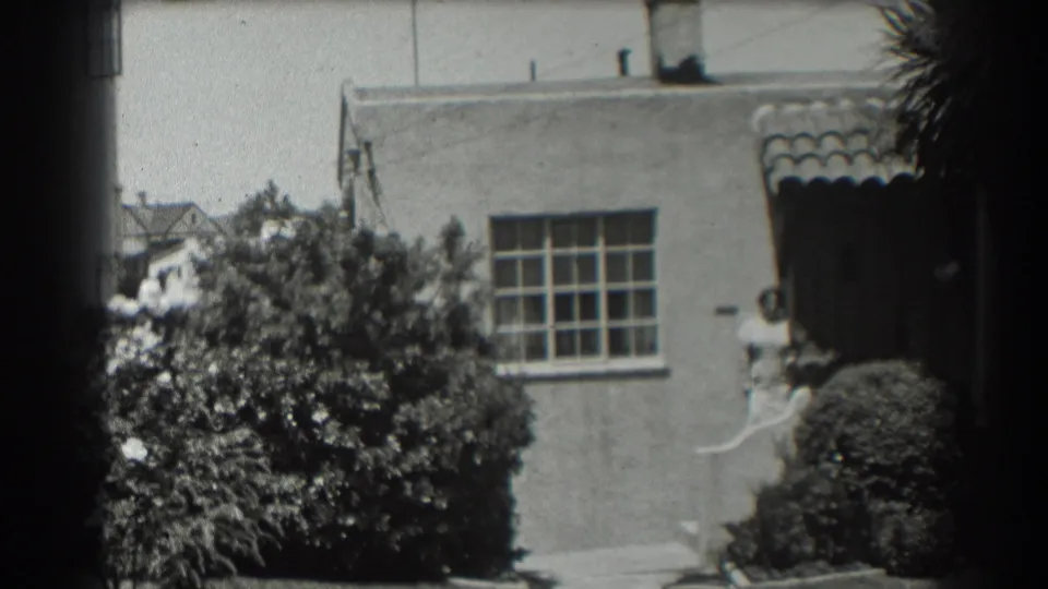 1939 San Francisco home-movie frame of a residential house exterior with bushes, tiled roof detail, and a person by the entrance.