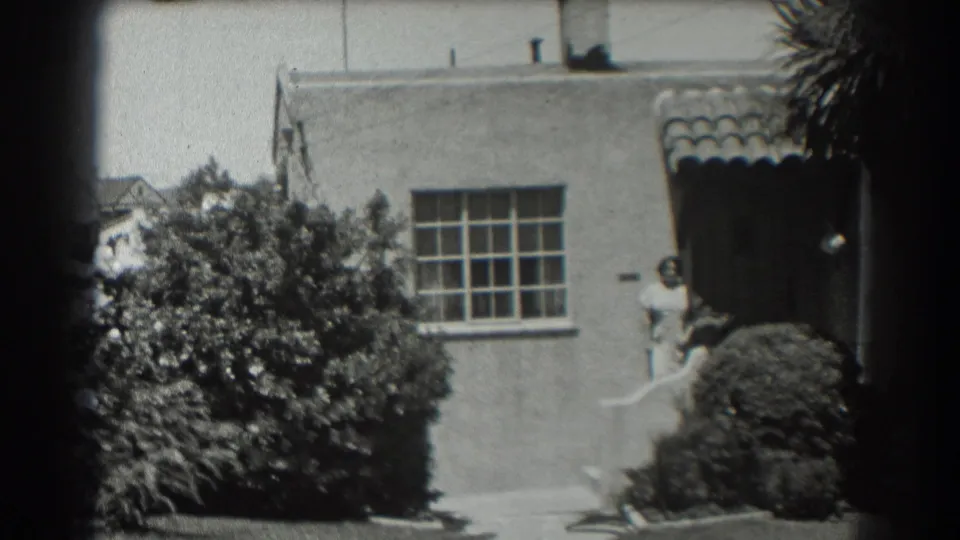 1939 San Francisco home-movie frame showing a stucco house exterior with shrubs, a tiled entry awning, and a woman by the front door.