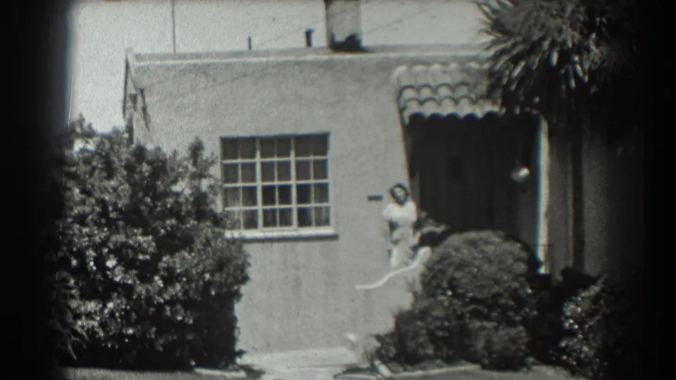 The pan centers the San Francisco house entrance, with palm fronds above, dense shrubs below, and a woman in light clothing near the door.