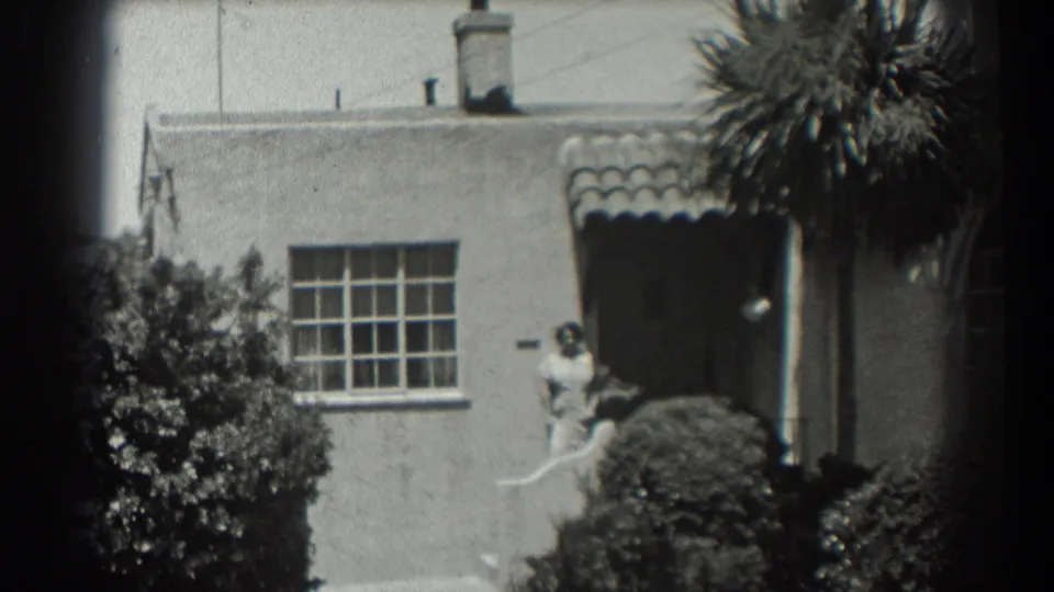 1939 home-movie frame of the stucco residence as the pan moves right, keeping the doorway, woman, palm, and garden shrubs in view.