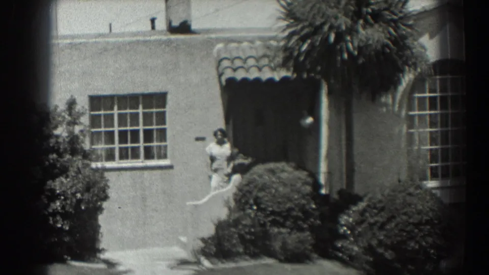 Near the end of the 1939 San Francisco pan, the woman remains near the door while the window, awning, palm fronds, and trimmed shrubs fill the frame.