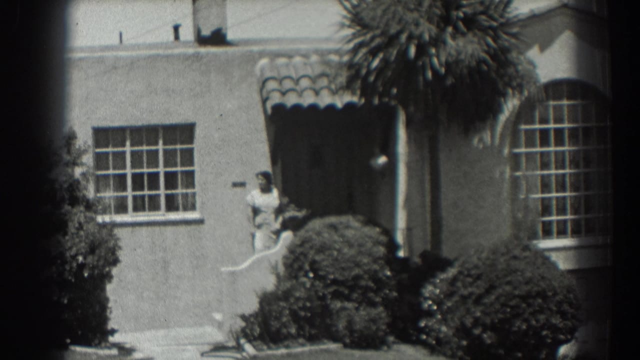 1939 San Francisco home-movie frame showing a woman near a stucco house doorway, with a tiled awning, shrubs, palm fronds, and divided windows.