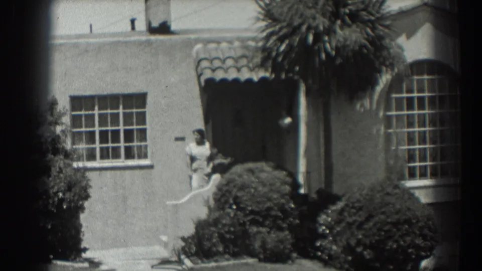 1939 San Francisco home-movie frame showing a woman near a stucco house doorway, with a tiled awning, shrubs, palm fronds, and divided windows.