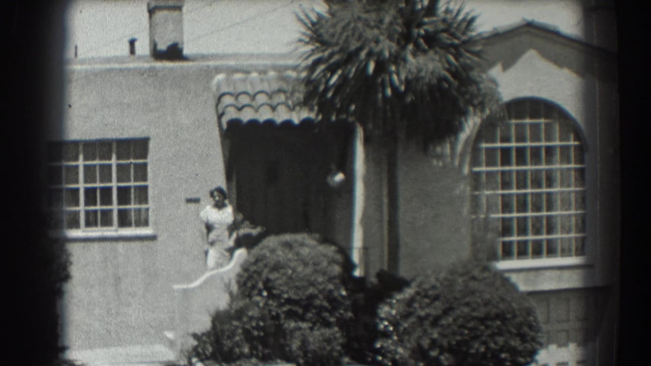 House front with a woman at the doorway, tiled awning, arched window, palm fronds, and shrubs.