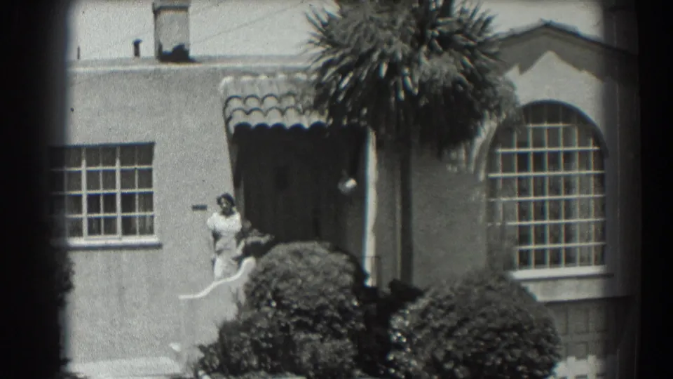 San Francisco 1939 residential exterior with a woman beside the entrance as the camera frames the tiled awning, arched window, shrubs, and palm.