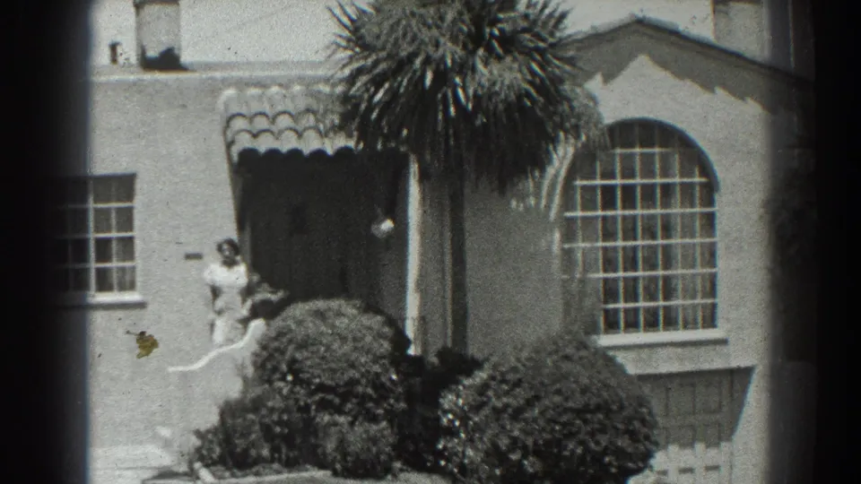 1939 San Francisco frame with the woman partly framed by shrubs near the doorway, while the pan emphasizes the tiled awning and arched window.