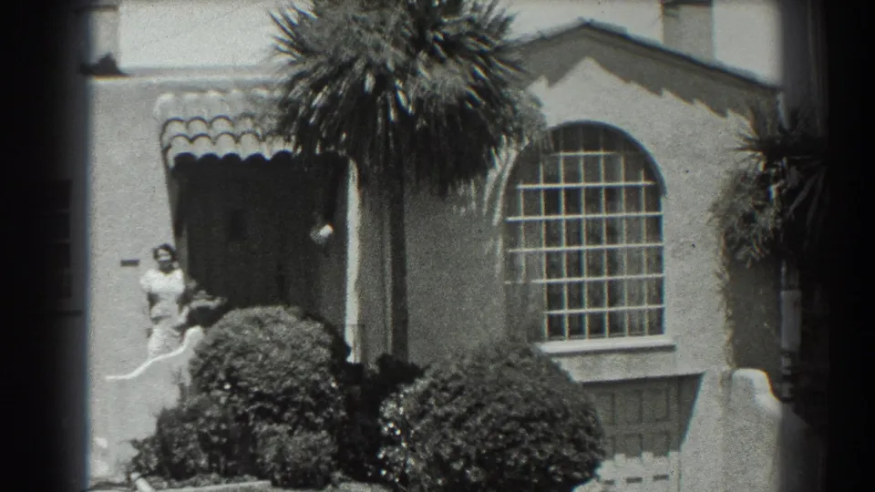 Late-1930s San Francisco home-movie frame showing the stucco house front, arched divided window, tiled awning, dense shrubs, palm, and doorway figure.