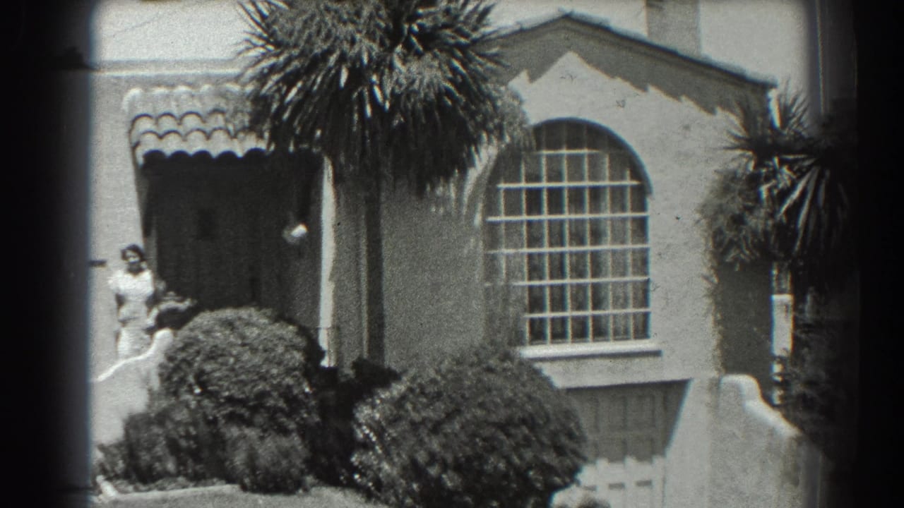 1939 San Francisco home-movie frame showing a woman near a stucco house entrance, with palms, shrubs, tiled roof edge, and an arched window.