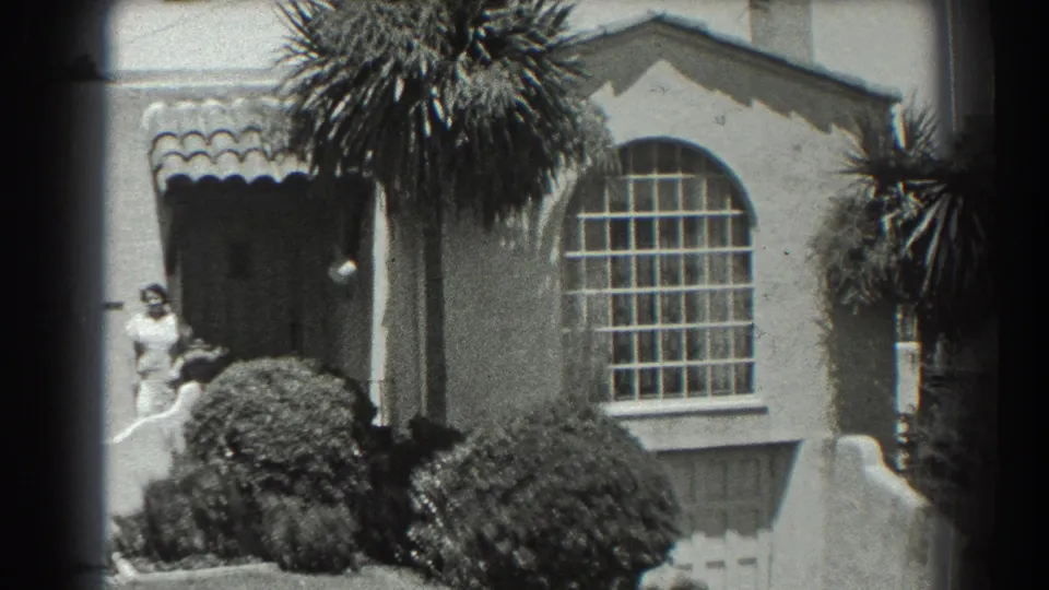 1939 San Francisco home-movie frame showing a woman near a stucco house entrance, with palms, shrubs, tiled roof edge, and an arched window.