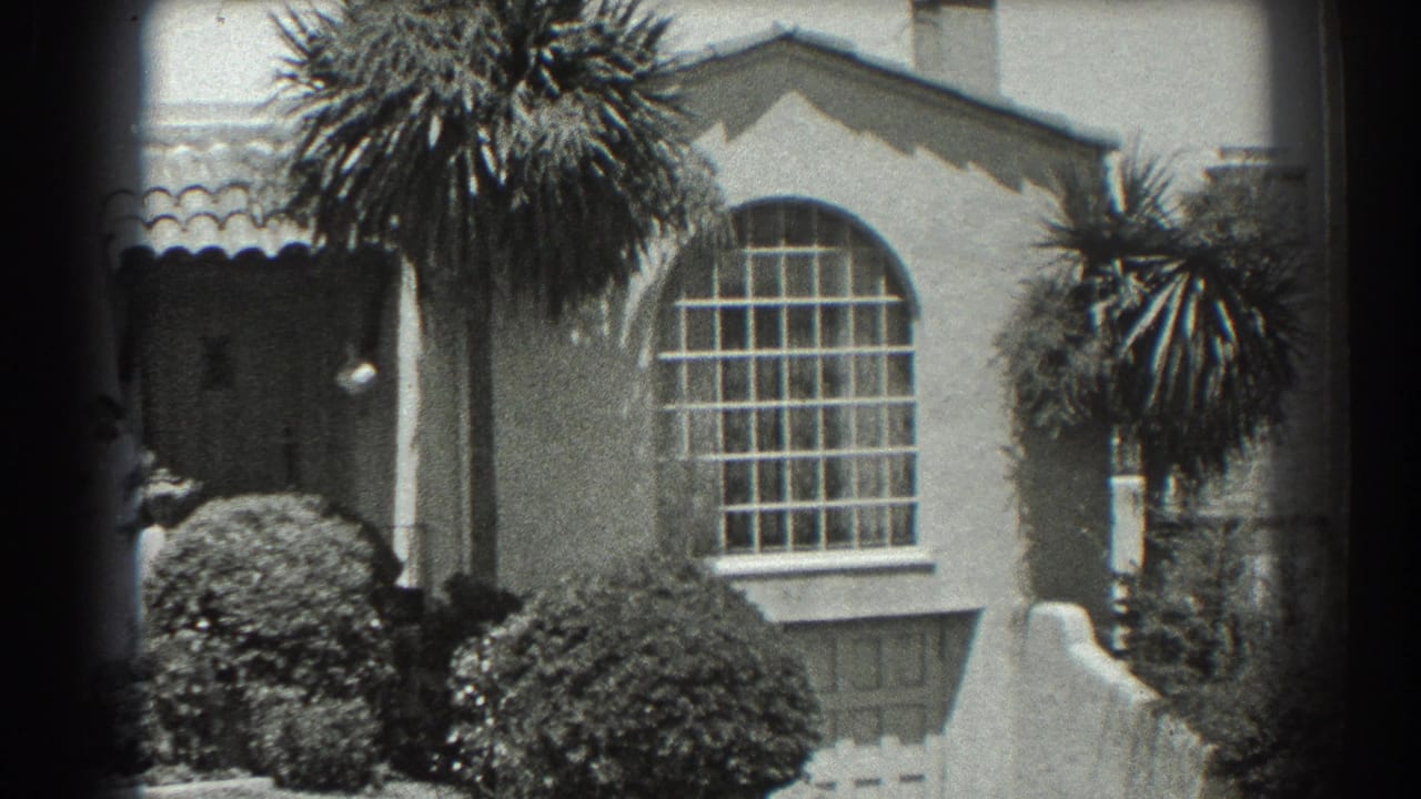 Stucco house exterior with arched window, palm trees, shrubs, and a woman partly hidden at left.