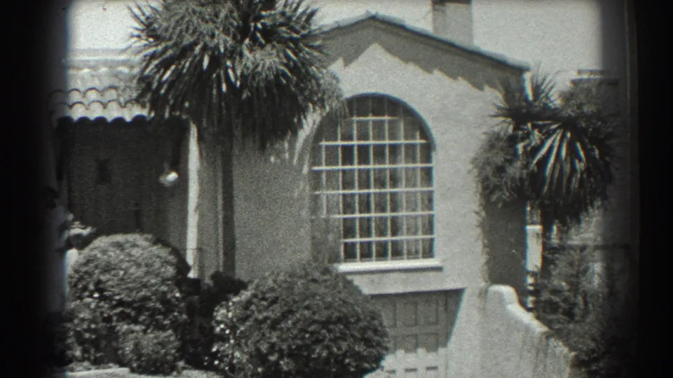 1939 San Francisco residential exterior with a woman partly visible by the doorway, an arched window, palms, tiled roof edge, and manicured shrubs.