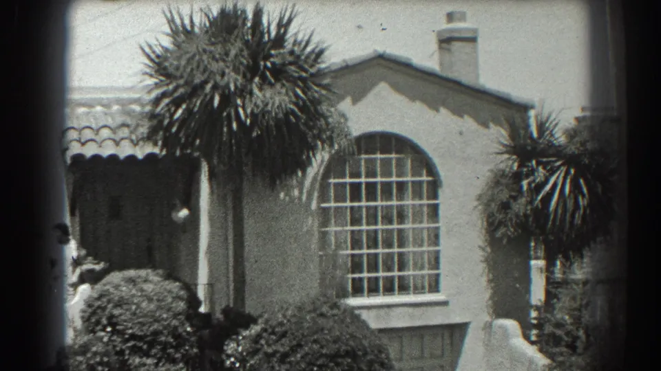 1939 San Francisco frame of a stucco residence with palms and shrubs, while a person is partly visible near the shaded entrance at left.