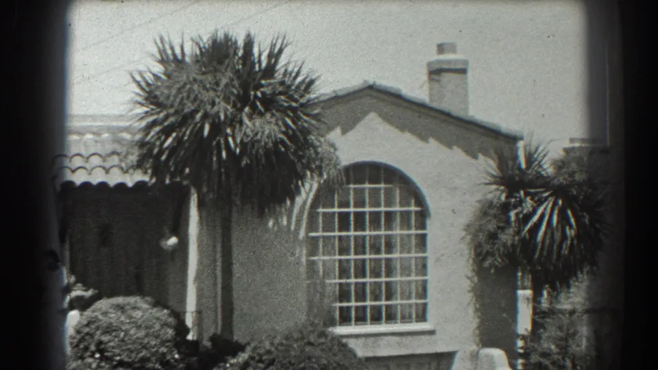 1939 San Francisco home-movie view of a stucco house exterior with a large palm, arched window, shrubs, and a partly seen person near the entry.