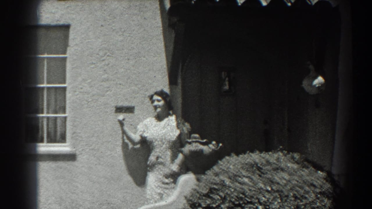 Woman in patterned dress gestures beside a stucco house entry and window.