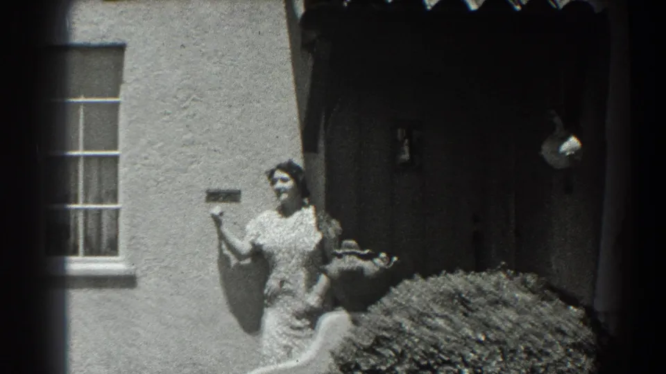 In 1939 San Francisco home-movie footage, a woman gestures outside a stucco house with a window, shaded doorway, shrub, and curved garden feature.