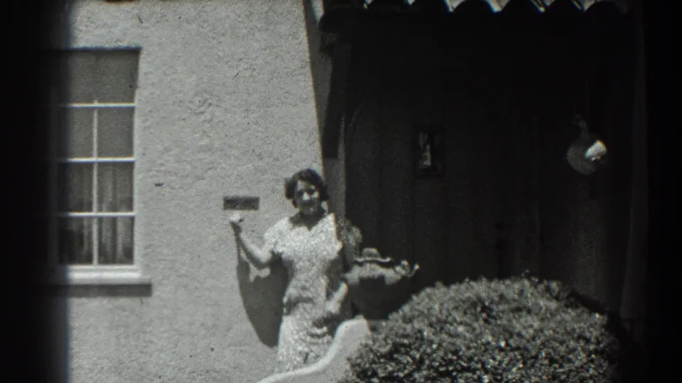 The woman stands outside the San Francisco stucco house, appearing to speak toward the camera near the arched entry and front shrub.