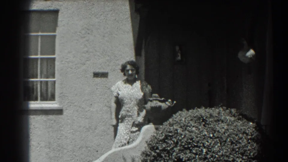 A woman in a patterned dress stands in bright sunlight outside the 1939 San Francisco residence, framed by a window, entry shadow, and shrub.