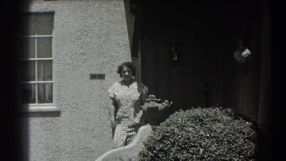 The final frame holds on the woman outside the stucco San Francisco house, with the dark entrance, window, shrub, and garden feature visible.