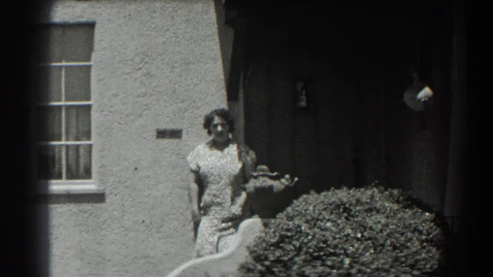 In supported 1939 San Francisco home-movie footage, a woman stands near a stucco house entry beside a window and shrub.