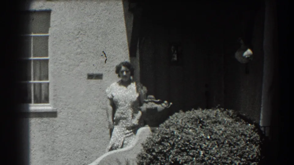 A woman stands outside a stucco residence in the 1939 San Francisco clip, with a doorway, window, and shrub behind her.