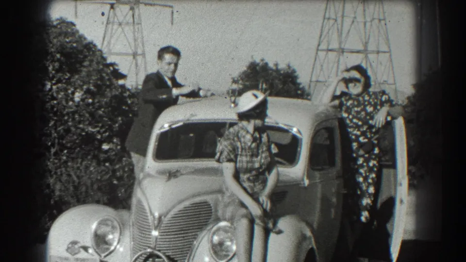 In the 1939 San Francisco home movie, two women and a man pose around a rounded prewar car near a residential exterior.