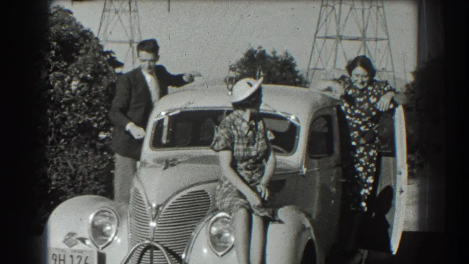 In 1939 San Francisco home-movie footage, three people pose around a classic sedan with trees and towers behind them.