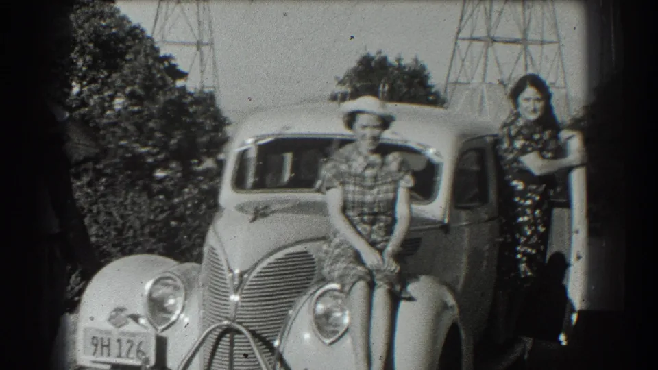 A black-and-white 8mm frame shows a younger female seated on the sedan fender and a woman leaning near the open door.