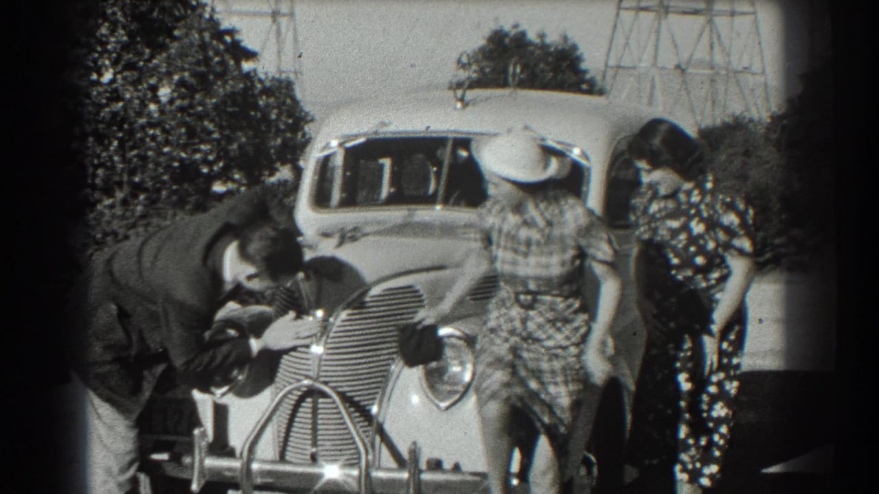 Woman in a plaid dress moves from the car hood as a man works near the front.