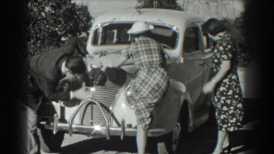 The plaid-dress woman turns toward the driver's side as another woman stands beside the car and the man remains near the hood.