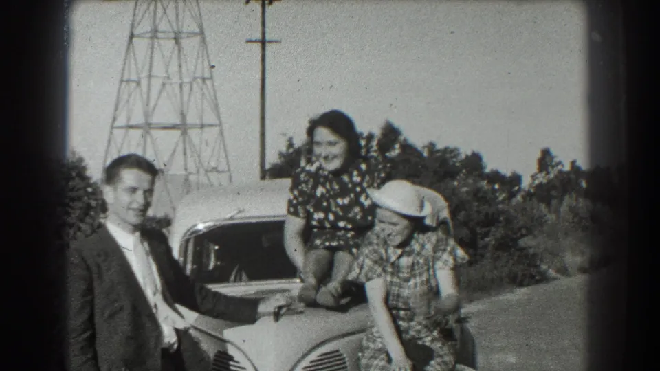 Three adults pose around the hood of the car, with two women leaning on or sitting near it and utility poles visible behind them.