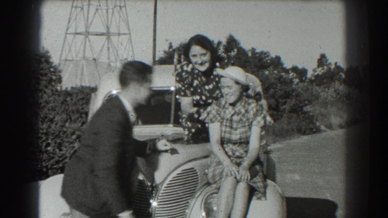 Man near a rounded car photographs or directs two women posing on the front of the vehicle.