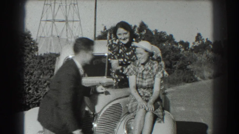 In a 1939 San Francisco home-movie frame, two women pose on a rounded car while a man leans in with a camera or gesture.