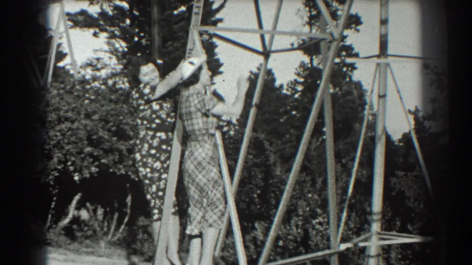 A woman in a plaid dress and hat climbs a metal pylon or ladder in the San Francisco home-movie footage as another woman watches.
