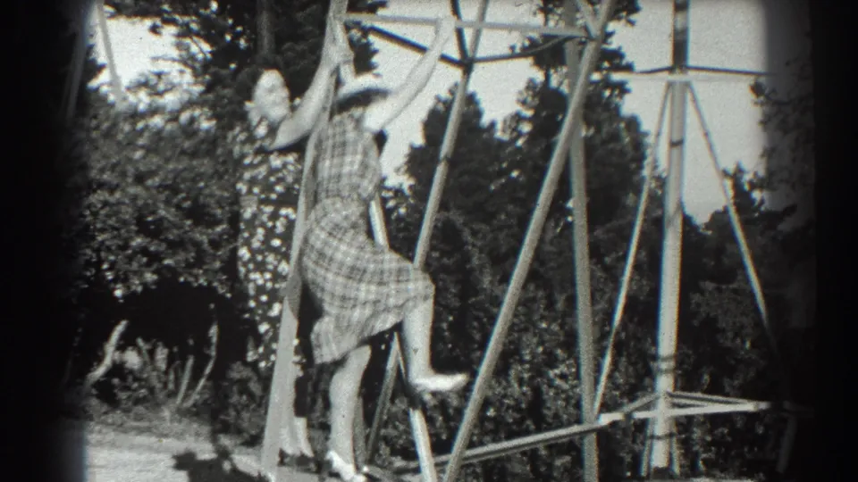The plaid-dressed woman continues climbing or posing on the metal structure, reaching up as another figure is partly hidden behind her.