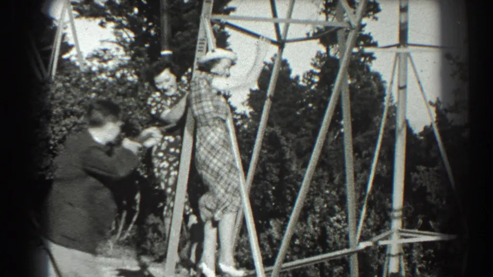 A man crouches or leans toward the woman on the metal pylon, apparently photographing or directing her pose in the 1939 outdoor scene.