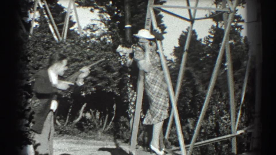 1939 San Francisco home-movie frame of a suited man playfully aiming a toy gun at a woman in a plaid dress and white hat outdoors.
