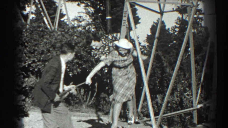 In a 1939 San Francisco backyard scene, a suited man crouches with a toy gun while a woman in a plaid dress reaches toward him.