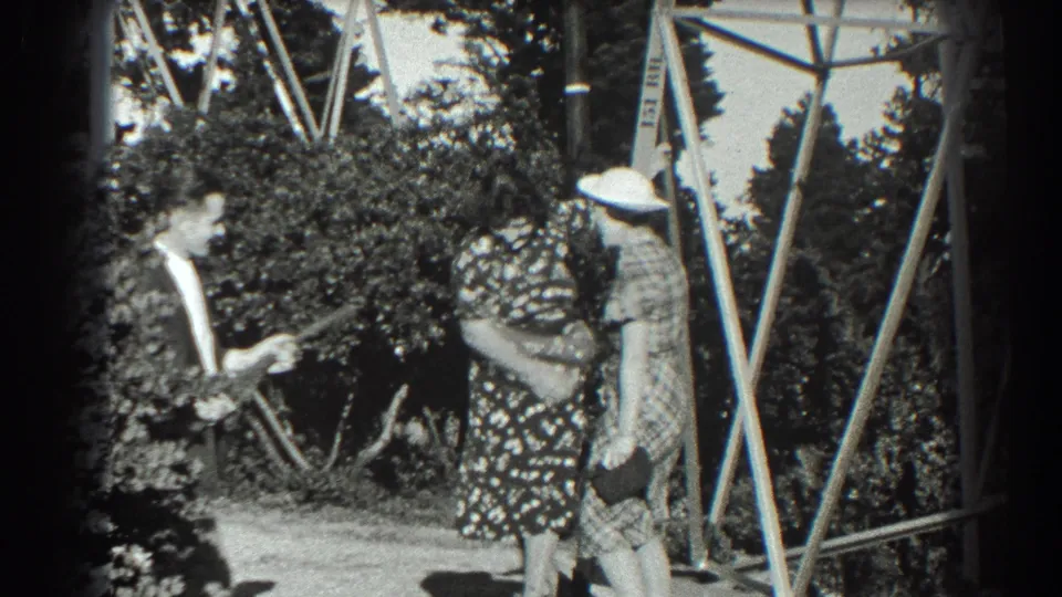 1939 San Francisco home-movie frame with a suited man holding a toy gun near two women in patterned dresses in a garden setting.