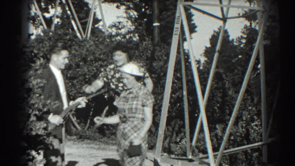 A 1939 San Francisco backyard home-movie frame shows two women smiling near a wooden structure while a suited man holds a toy gun.