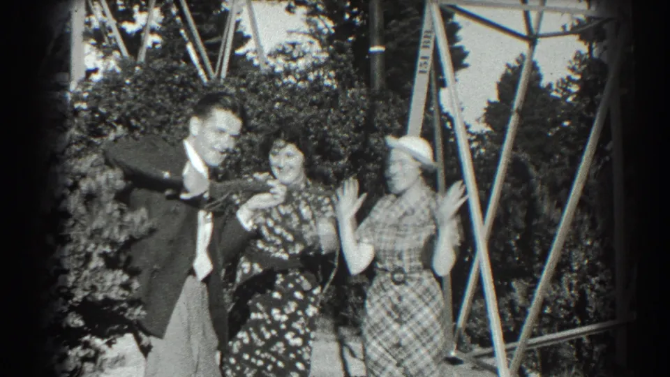 A close playful moment shows the suited man smiling beside two women with raised hands, with foliage and metal supports behind them.