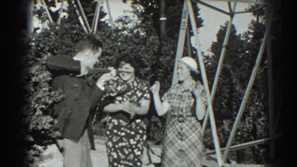 In 1939 San Francisco home-movie footage, a suited man and two women in patterned dresses pose playfully near a metal garden trellis.