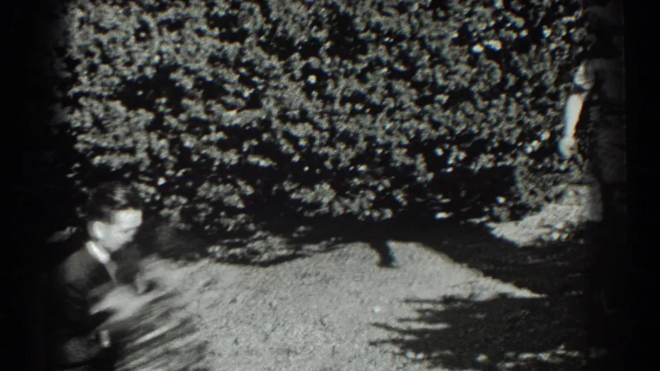 A man in a dark suit crosses a sunlit garden area, partly blurred by motion, with dense shrubs behind him.